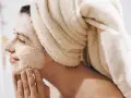 Frau mit Handtuch auf dem Kopf trägt Gesichtsmaske und massiert sie sanft ein.