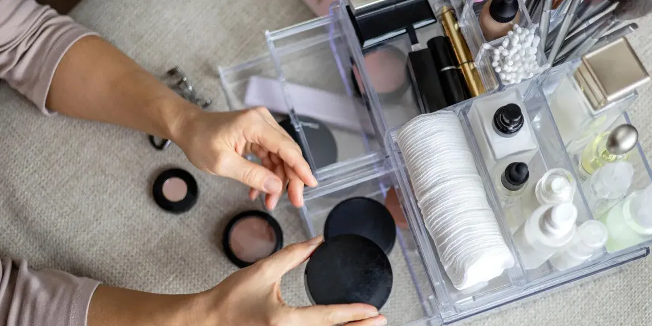 Hände sortieren Kosmetik in einem transparenten Organizer mit Pinseln, Puderdosen und Cremes.