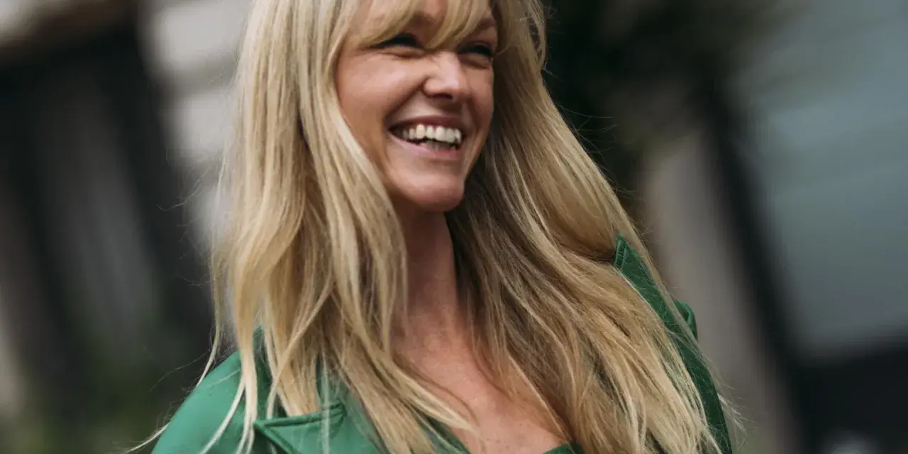 Frau mit langen blonden Haaren und Pony, die lacht und ein grünes Jackett trägt.