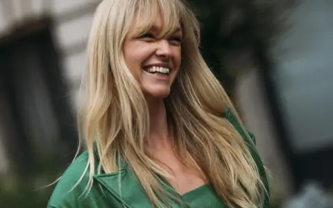 Frau mit langen blonden Haaren und Pony, die lacht und ein grünes Jackett trägt.