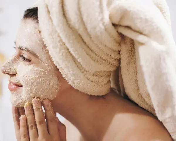 Frau mit Handtuch auf dem Kopf trägt Gesichtsmaske und massiert sie sanft ein.