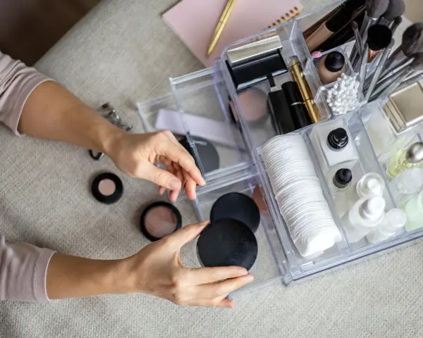 Hände sortieren Kosmetik in einem transparenten Organizer mit Pinseln, Puderdosen und Cremes.