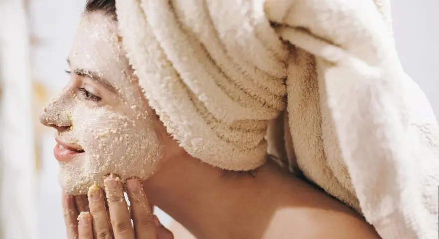Frau mit Handtuch auf dem Kopf trägt Gesichtsmaske und massiert sie sanft ein.