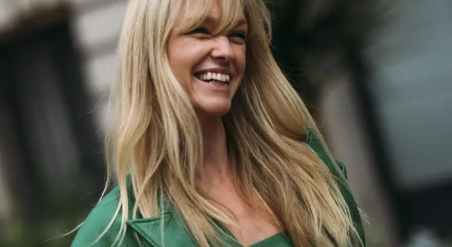 Frau mit langen blonden Haaren und Pony, die lacht und ein grünes Jackett trägt.